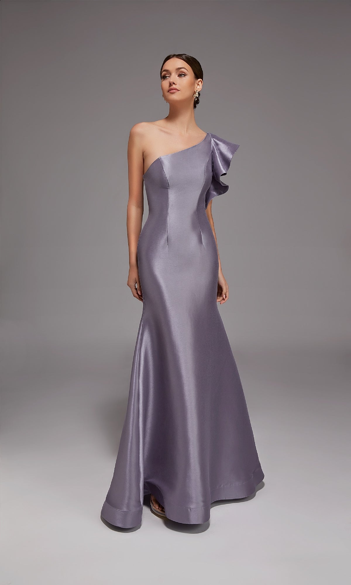 A woman stands against a plain gray backdrop wearing the Alyce 27826 long formal dress, a floor-length, one-shoulder metallic lavender gown with ruffled shoulder detail.